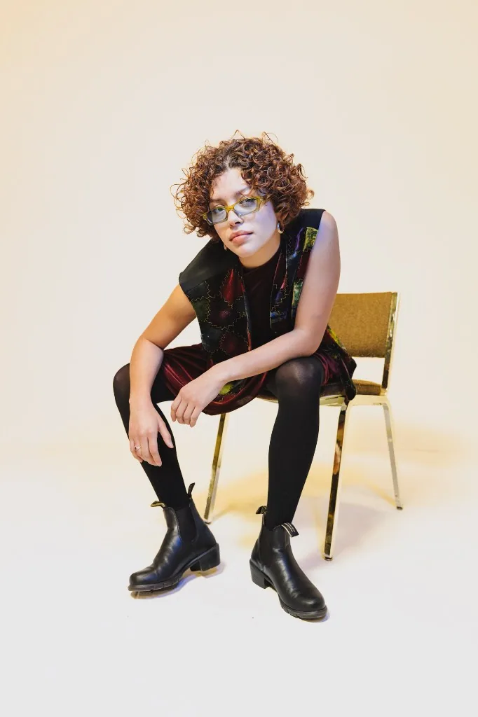 Studio portrait seated on a vintage chair in a patchwork vest and dress.