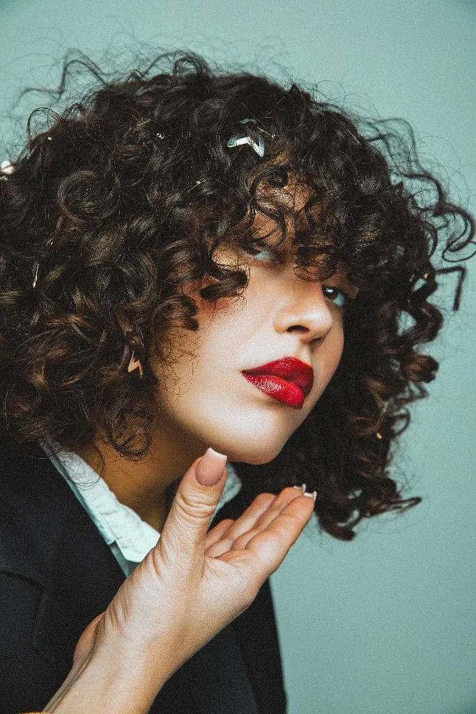 Artistic portrait with curly hair and bold red lipstick against a teal background.