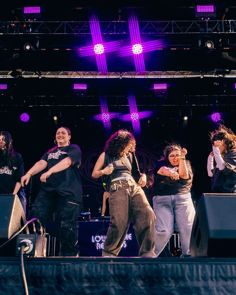 Group of performers on an outdoor stage under purple concert lighting.