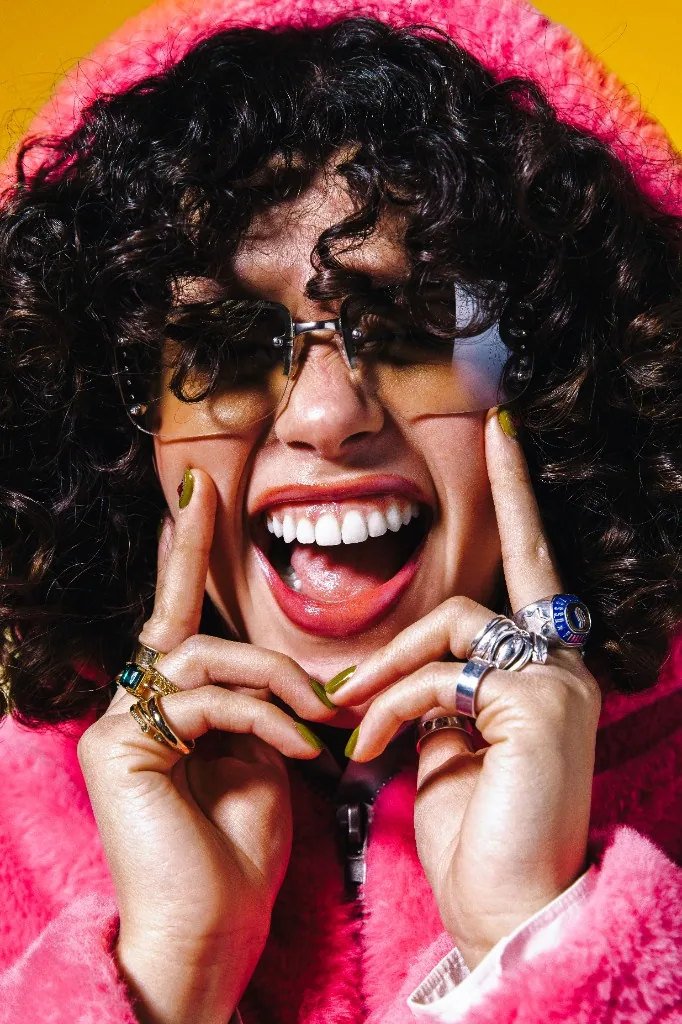 Close-up portrait in a pink jacket and yellow sunglasses against a bright yellow backdrop.